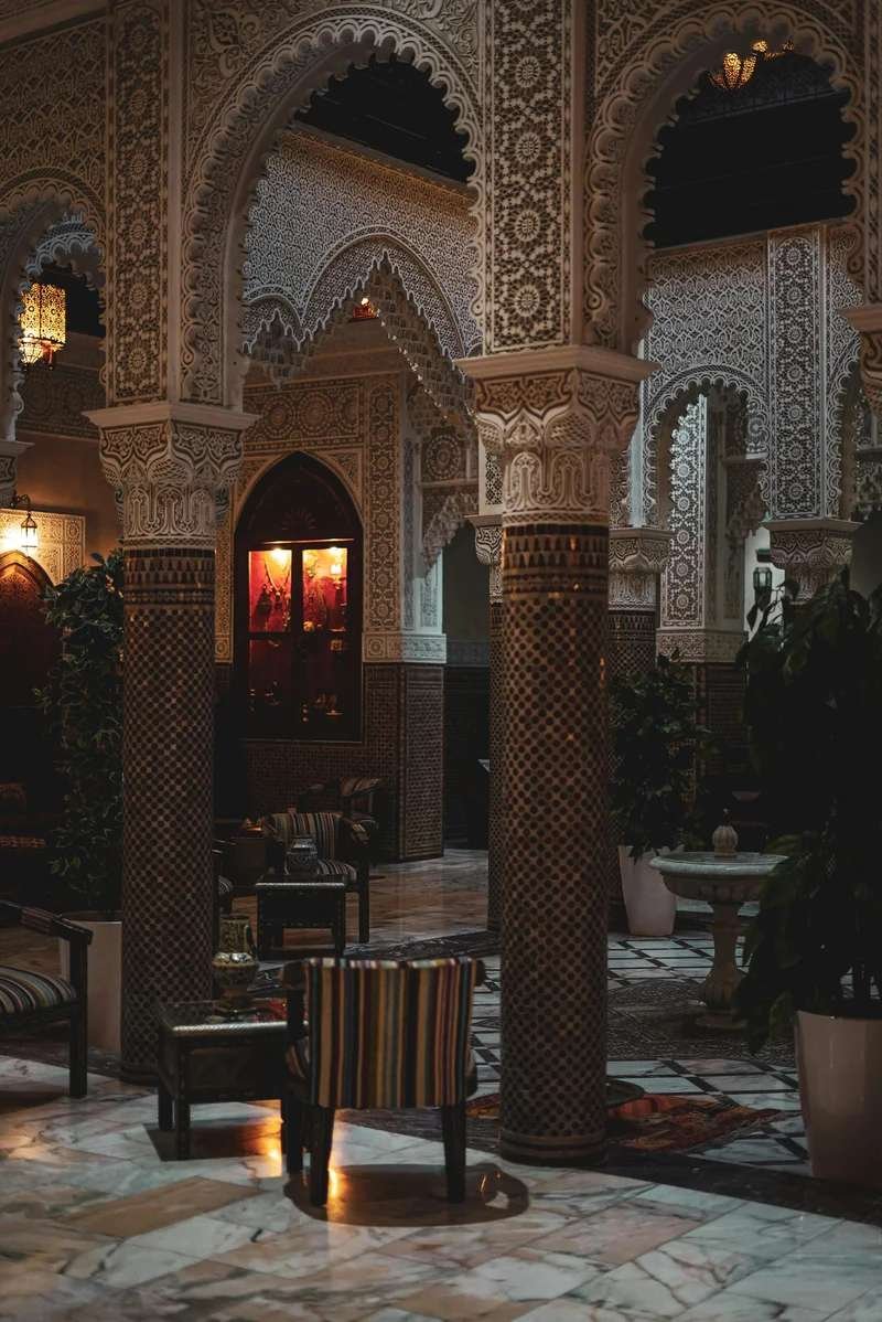 Exquisite traditional Moroccan riad interior featuring elaborate stucco carvings, horseshoe archways, ornate zellige columns, ambient brass lanterns, and authentic Islamic geometric patterns demonstrating supreme craftsmanship