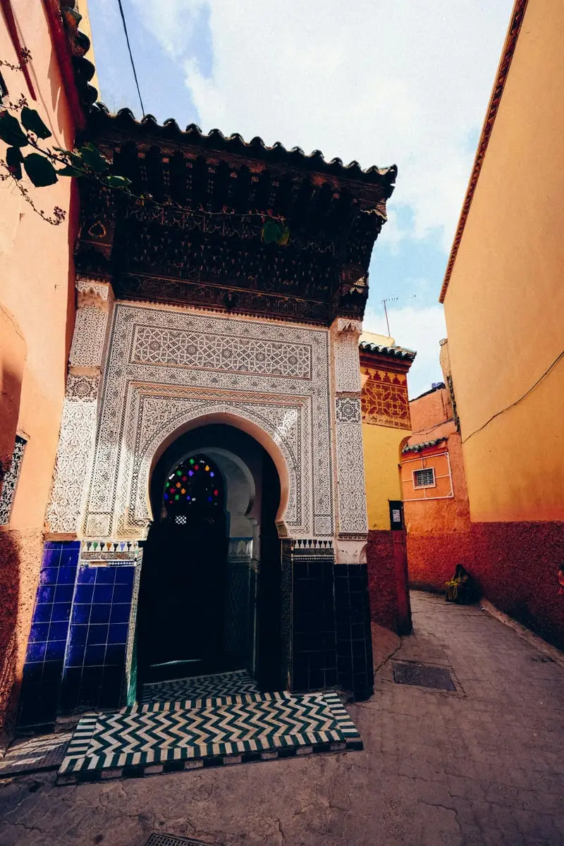 Stunning traditional Moroccan entrance gate with intricate Islamic geometric tilework, carved wooden zellige details, colorful stained glass window, and chevron-patterned mosaic flooring in the historic medina