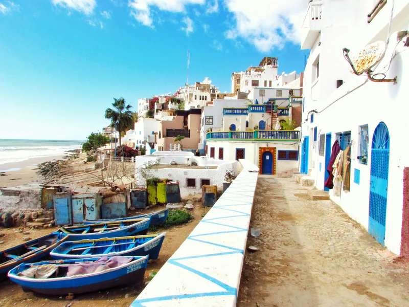Historic coastal architecture with whitewashed buildings and traditional blue fishing boats along Asilah's seafront, reflecting the atmospheric rampart-lined shores of Morocco's Atlantic coast