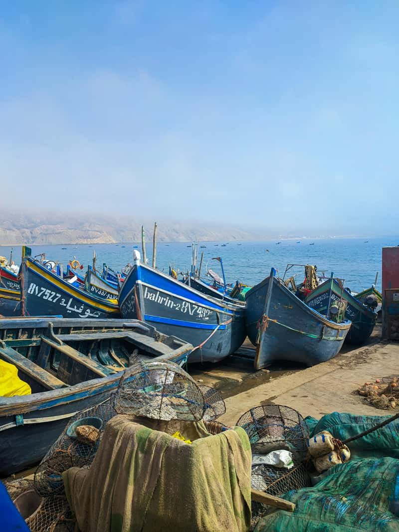 Blue wooden fishing boats hauled up on the quayside at Agadir's port, with nets and traps stacked in the foreground and a headland fading into haze across the bay.
