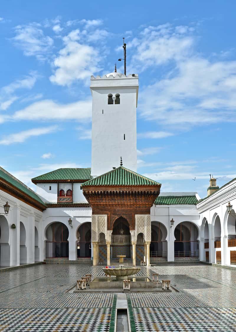 Courtyard of the Al-Qarawiyyin mosque in Fes, with white arcades, green-tiled roofs and a carved cedar pavilion sheltering the fountain