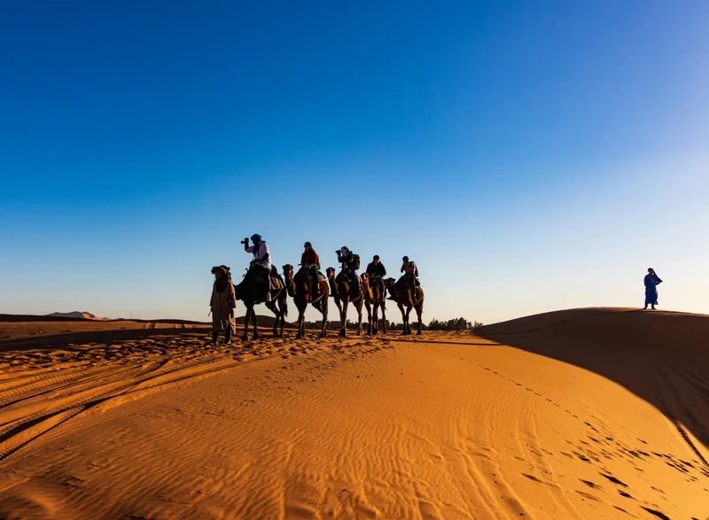 Camel caravan trekking across golden Sahara desert dunes during sunset with Berber guides