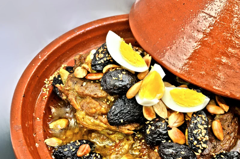 Close-up of traditional Moroccan lamb tagine garnished with prunes, toasted almonds, sesame seeds, and hard-boiled eggs in clay pot
