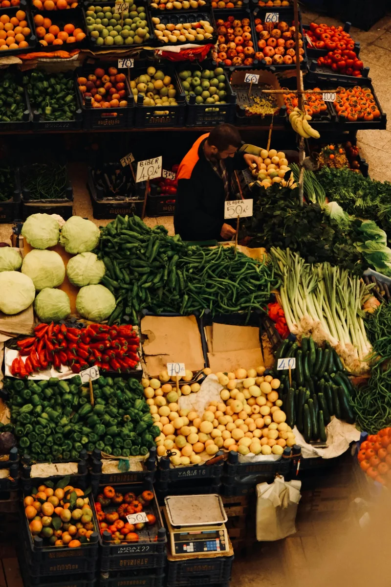 Penjual pasar Maroko autentik dikelilingi oleh sayuran segar berlimpah dan buah berwarna termasuk paprika, kembang kol, tomat, dan jeruk menunjukkan budaya makanan lokal yang semarak dan kehidupan sehari-hari